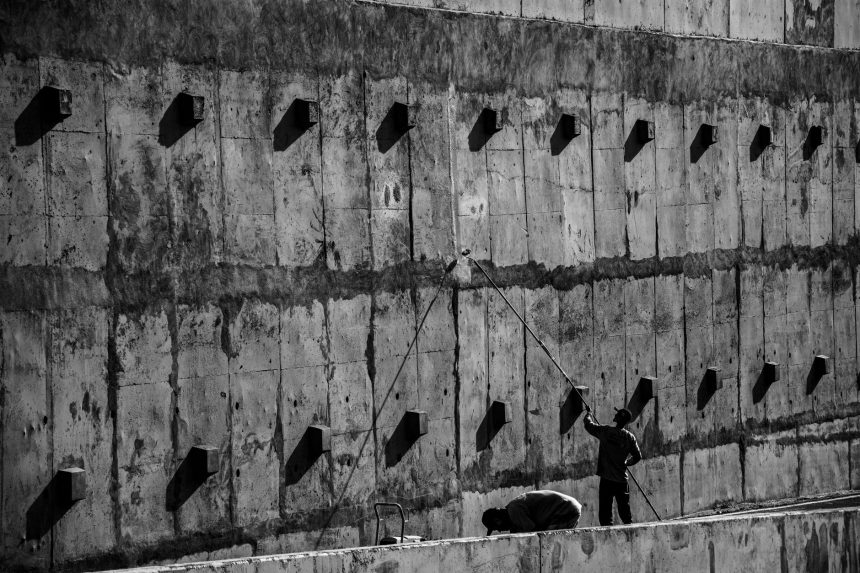 Dois trabalhadores pintam uma grande parede de concreto em um ambiente de trabalho.