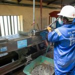 Trabalhador usando uniforme azul opera uma mesa vibratória para processamento de minério em uma unidade industrial.
